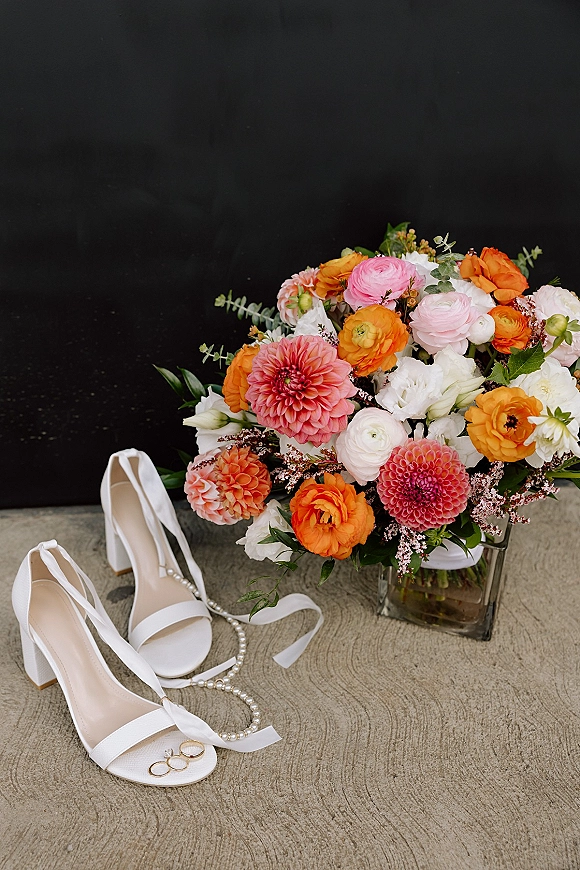 Wedding details flat lay with bridal shoes flat lay, bouquet in a glass vase, rings, pearl necklace and ribbon on black concrete backdrop