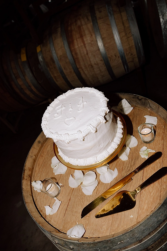 Wedding cake on a gold cake stand with piped buttercream icing, cake knife and server, votive candles and rose petals on a wood barrel table