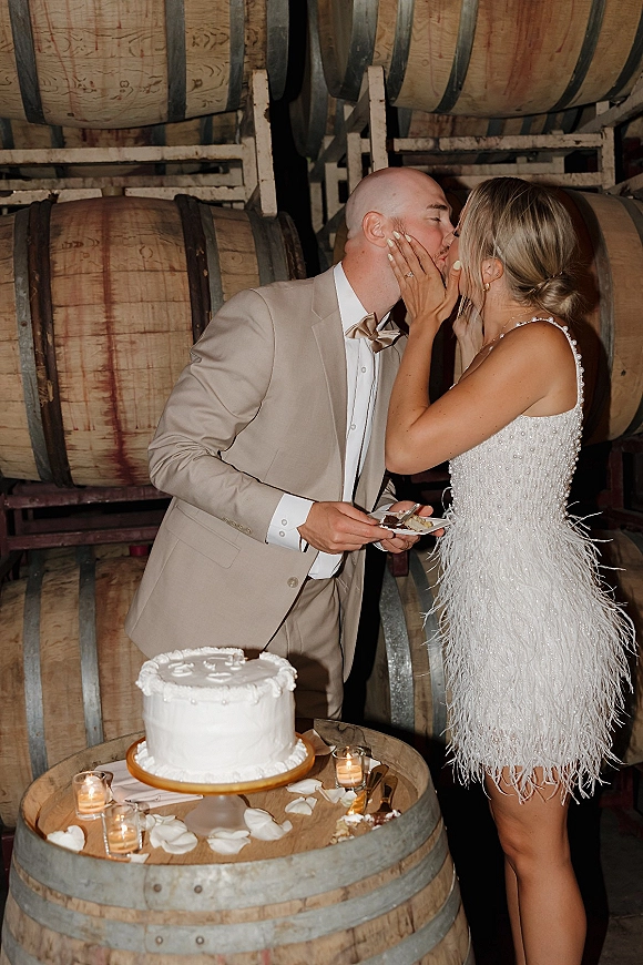 Wedding cake cutting as bride in a beaded fringe dress and groom in a beige suit share a bite beside wine barrels, rose petals, and candles