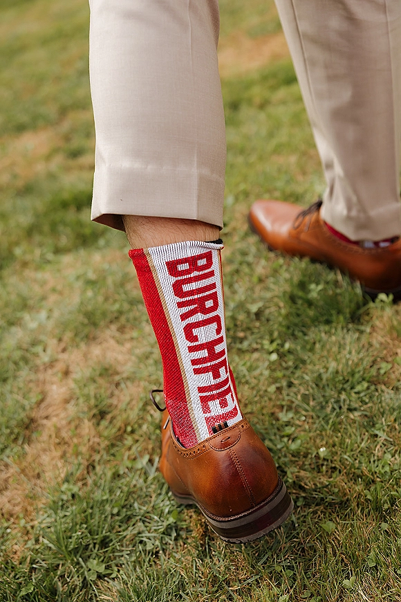 Groom socks with funny groom socks peek above brown leather dress shoes and tan suit pants, standing on a grass lawn outdoors