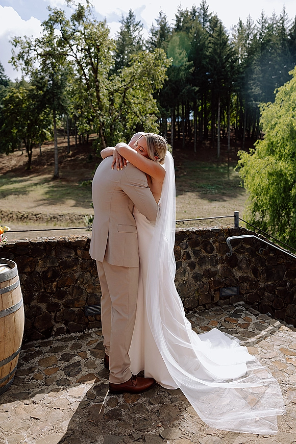 Wedding couple hug as bride and groom embrace, her long veil train flowing behind on a stone patio by a rustic wall and meadow