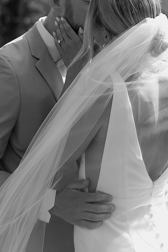 Wedding kiss portrait in black and white, bride and groom kissing under a veil, her ringed hand on his face with greenery behind