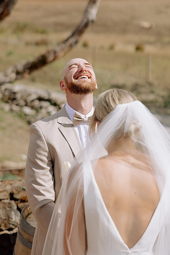 First look moment as bride in a long veil and open-back dress approaches groom in a beige suit by a stone wall on a hillside outdoors