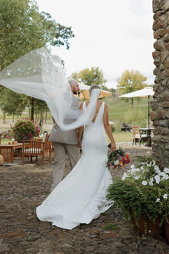 Couple portrait of bride and groom walking away holding hands, long veil blowing over stone patio with umbrellas and hills behind