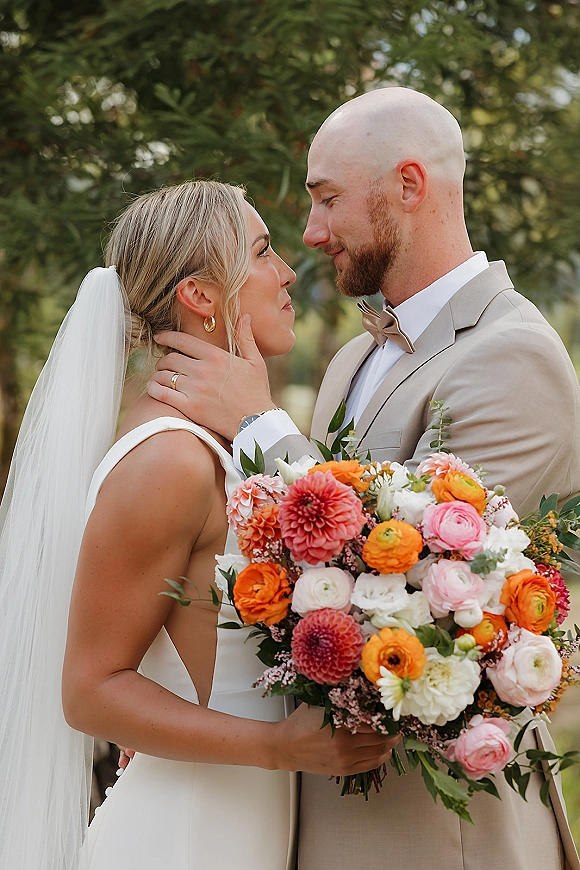 Couple portrait of bride and groom close up, groom hand on bride face as she holds a bright bouquet with veil amid trees
