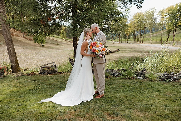 Couple portrait of bride and groom embracing with a forehead kiss, bride holding a colorful bouquet and long veil under a tree in a vineyard lawn.