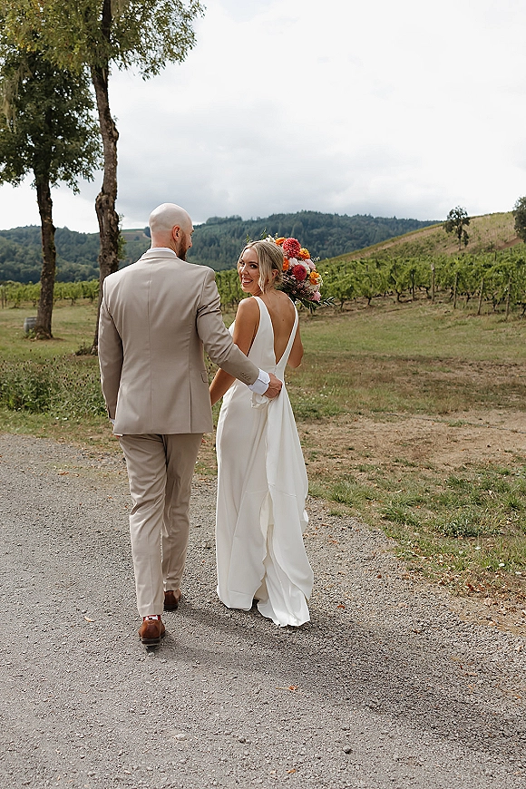 Couple portrait of bride and groom walking away as she looks over her shoulder, holding a bouquet on a vineyard gravel path