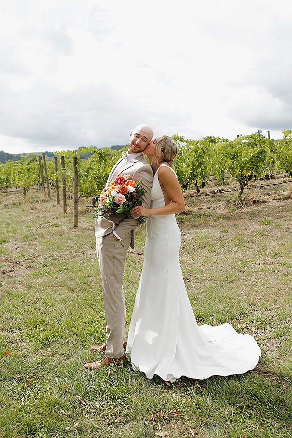 Couple portrait of bride kissing groom, bride holding a colorful bouquet beside a vineyard with grass field and cloudy sky backdrop