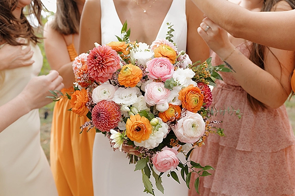 Bridal bouquet of dahlias and ranunculus in coral and pink with lush greenery, held by the bride as the wedding party blurs behind.