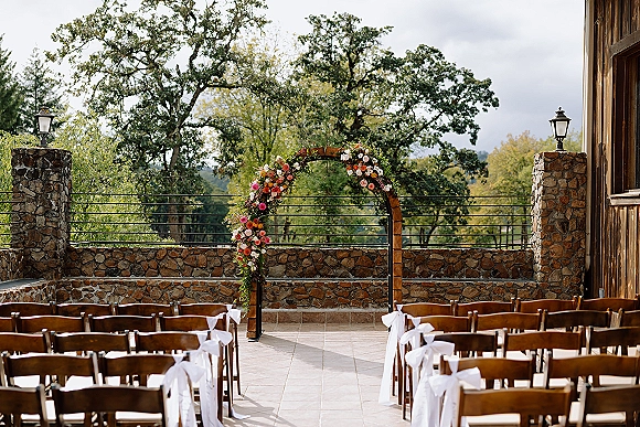 Ceremony setup for an outdoor wedding ceremony with a floral arch over a wooden frame, white chair bows lining a patio by stone wall