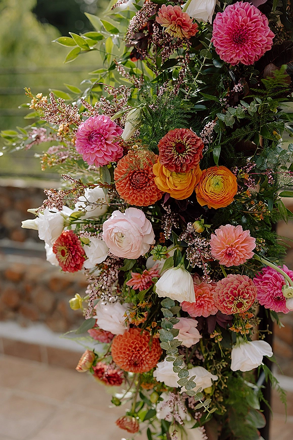 Wedding floral arrangement with pink and orange blooms and eucalyptus accents beside a garden railing and stone wall outdoors