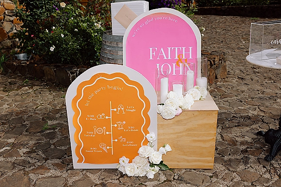 Wedding welcome sign on a wooden box pedestal with acrylic signage and white roses, pillar candles in hurricanes on a stone patio wall backdrop