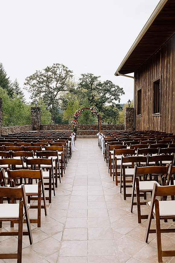 Ceremony setup with outdoor wedding ceremony seating, wood folding chairs lined with white ribbons facing a floral arch on a stone patio by a barn wall