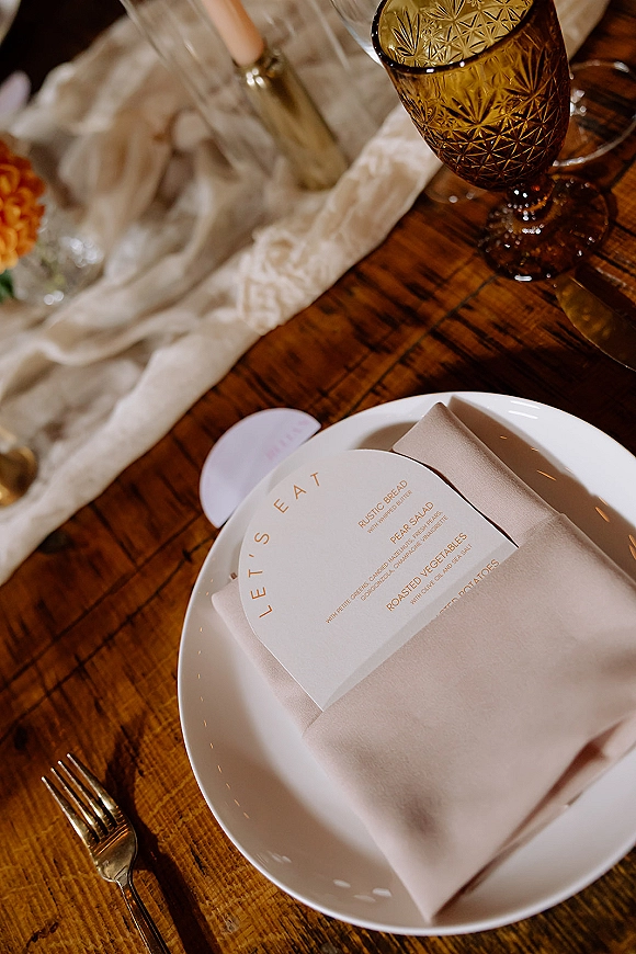 Wedding place setting with wedding menu card on a white plate, amber goblet, taper candle, and beige napkin on a rustic wood table
