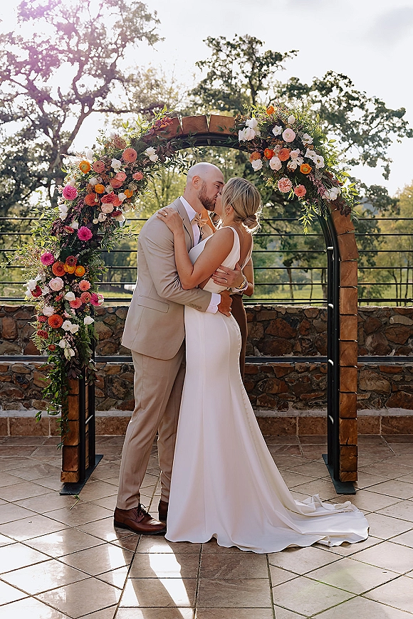 Wedding kiss portrait of bride and groom kissing beneath a colorful floral arch on an outdoor stone patio with trees and sky behind