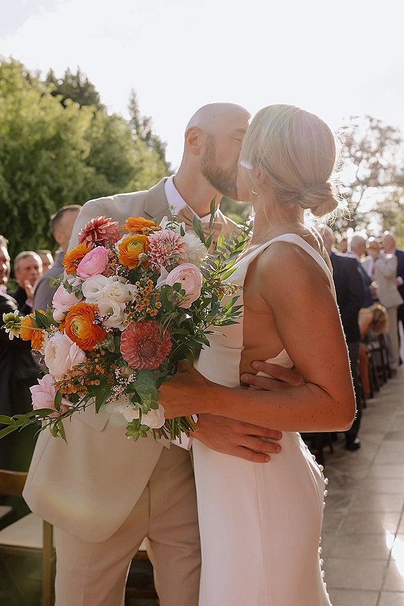 Wedding kiss as the bride and groom share their first kiss at ceremony, bouquet in hand, guests seated along a sunlit outdoor aisle