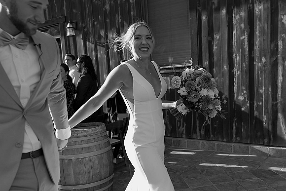 Newlywed couple portrait with bride holding bouquet, smiling as they hold hands on an outdoor patio by rustic wood siding and wine barrel