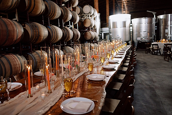 Reception tablescape with taper candles and amber goblets on a long wood banquet table, set in an industrial winery barrel room with tanks