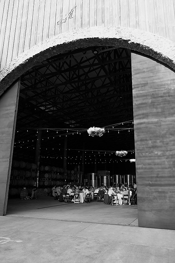 Reception setup with string lights and hanging floral chandeliers over banquet tables in an industrial warehouse, arched doorway and truss ceiling