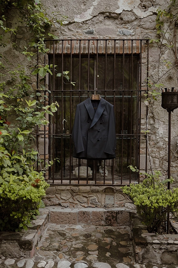 Groom suit flat lay with black wedding suit jacket on a wooden hanger by window bars against a vine-covered stone wall courtyard backdrop