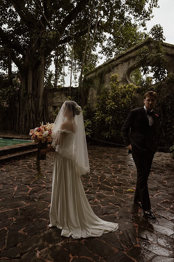 Wedding couple portrait with the bride holding bouquet beside her groom in tuxedo on a stone patio by a pool with tropical greenery