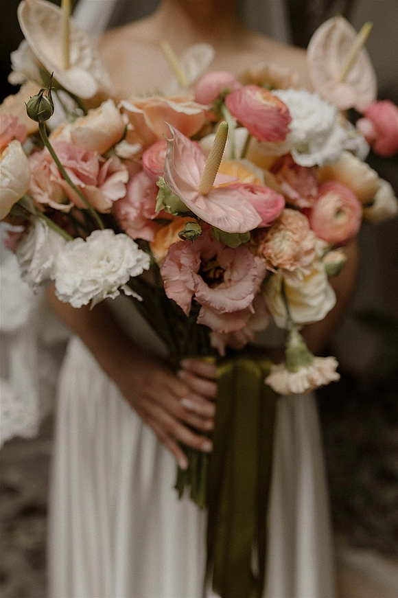 Bridal bouquet with anthurium wedding bouquet blooms, roses and ranunculus, tied with a long trailing ribbon in the bride’s hands indoors