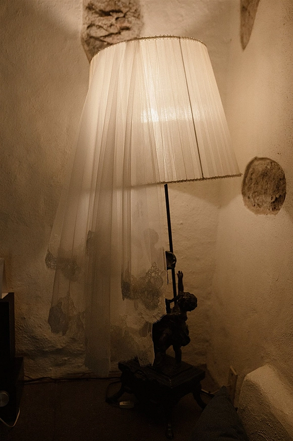 Bridal veil detail with lace edge veil draped over a vintage floor lamp, sheer tulle falling against a textured plaster wall in warm light