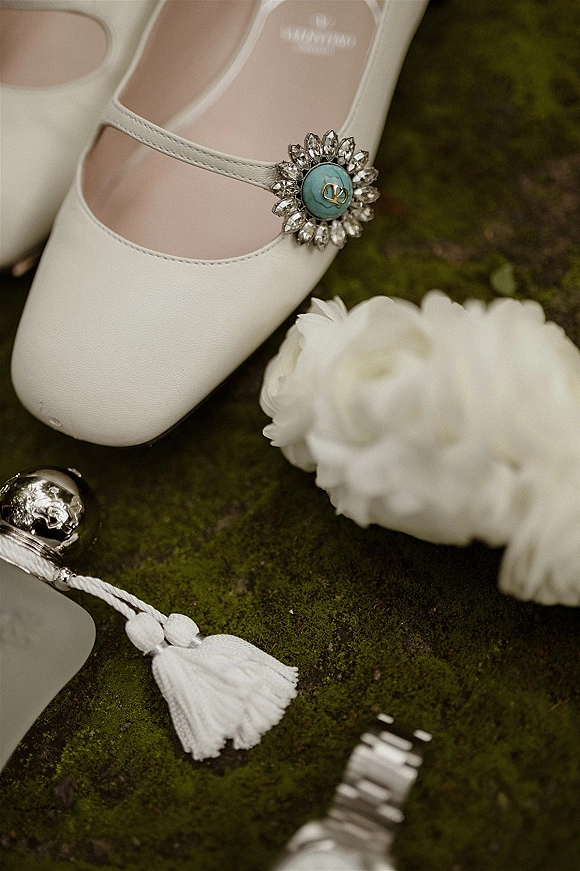 Bridal shoes in a wedding shoe flat lay, white Mary Jane heels with jeweled buckle and turquoise detail beside perfume on mossy surface