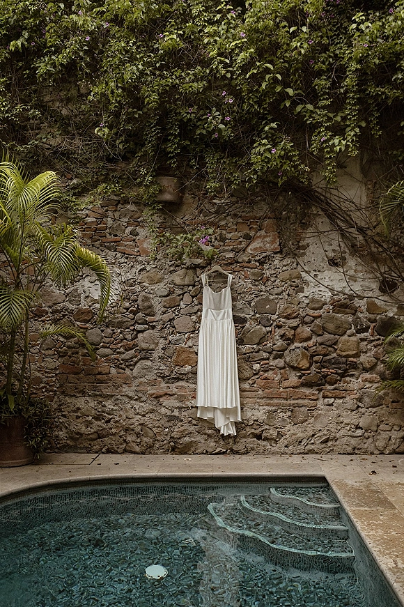 Wedding dress on hanger with a potted palm accent, hanging by a vine-covered stone wall beside the pool edge and water
