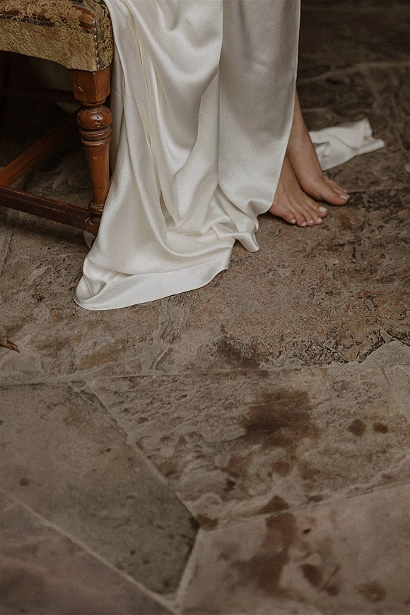 Wedding dress detail of a satin wedding dress hem and train draping and pooling beside a chair on a stone tile floor