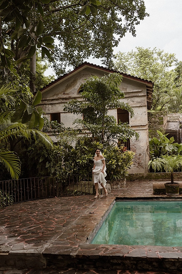 Bridal portrait of a bride in an off the shoulder wedding dress and veil, walking poolside in a tropical courtyard with stone patio