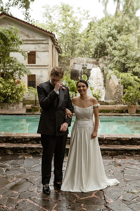 Couple portrait of bride holding the groom’s arm in a veil and off-the-shoulder dress beside a pool and waterfall backdrop
