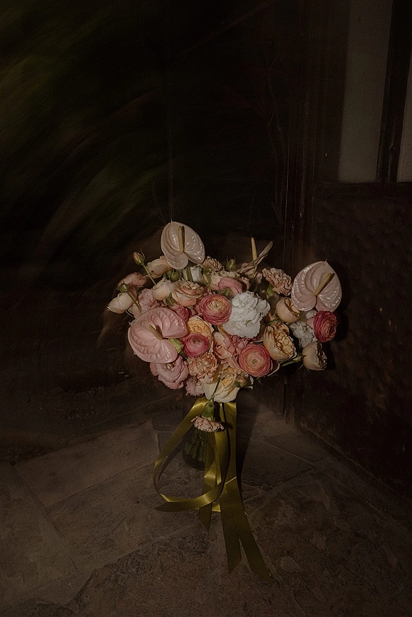 Bridal bouquet with anthurium wedding bouquet blooms, roses and ranunculus, finished with flowing ribbon tails against stone floor doorway backdrop