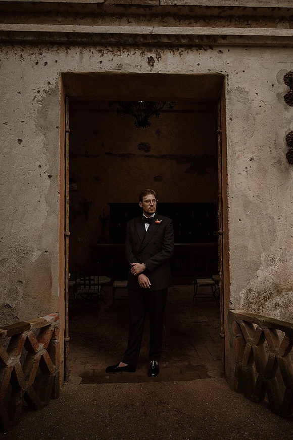 Groom portrait in a black tuxedo with bow tie, boutonniere, and eyeglasses standing in a stone doorway of a rustic interior with chandelier