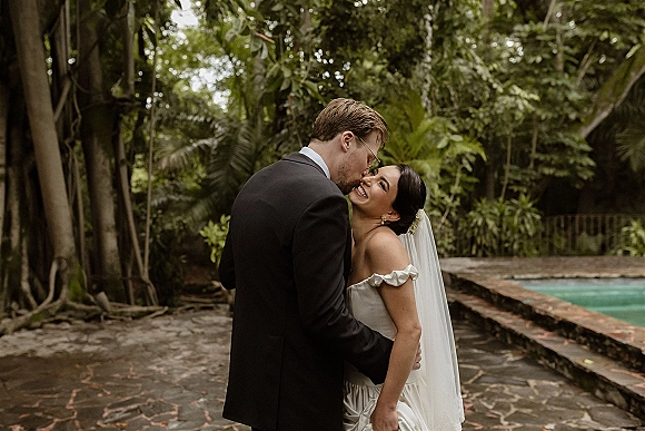 Couple portrait of bride and groom hug as he kisses her forehead beside a pool, her bridal veil draped over a strapless dress