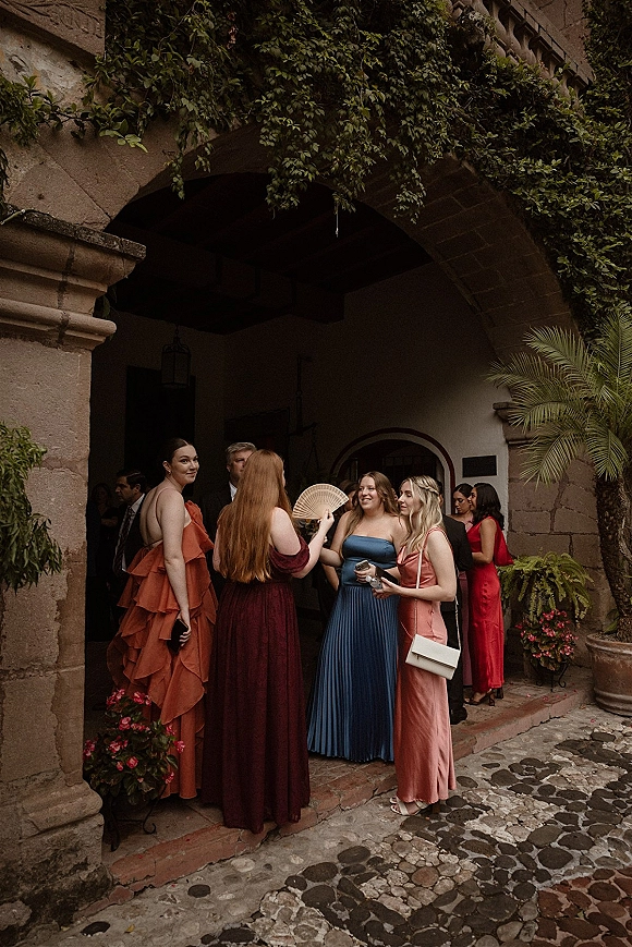 Wedding guests at wedding cocktail hour chat in colorful bridesmaid dresses under an ivy-covered stone archway in a cobblestone courtyard