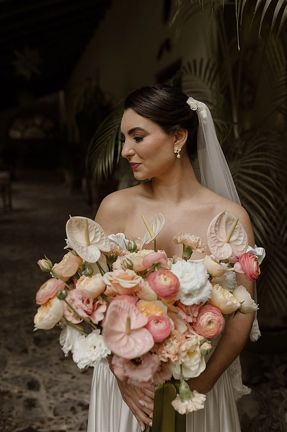 Bridal portrait of a bride holding bouquet with anthurium, peach blooms and ribbon, wearing veil and off-shoulder dress by tropical foliage