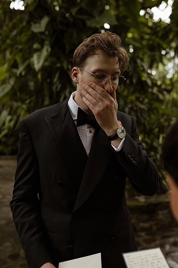 Groom portrait of a groom in tuxedo covering his mouth while reading vow letters, wearing glasses and boutonniere on a garden path