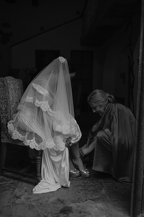 Getting ready moment as bride in a satin robe and lace-trimmed veil sits while someone helps with platform heels by a doorway