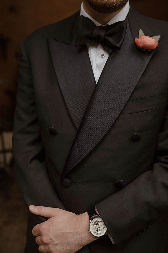 Groom tuxedo with black tie groom details, satin shawl lapel and black bow tie, peach rose boutonniere and wristwatch in warm interior