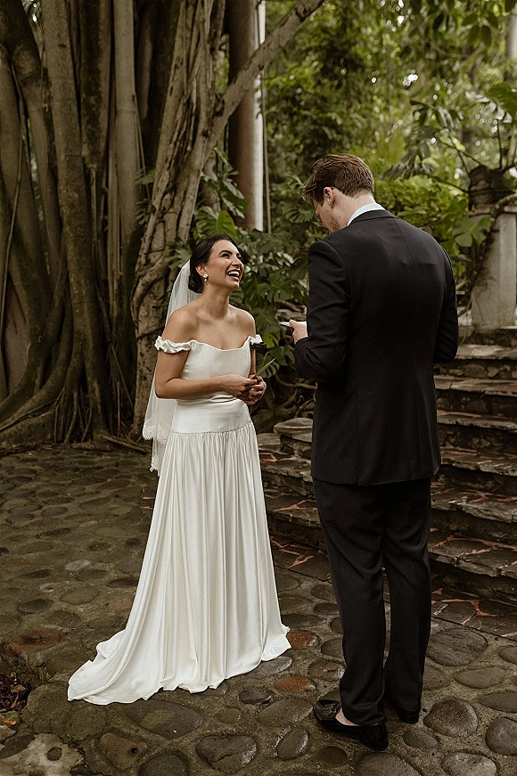 Wedding vows as groom reads to bride in an intimate vow exchange on stone steps, her off-the-shoulder gown and veil framed by greenery