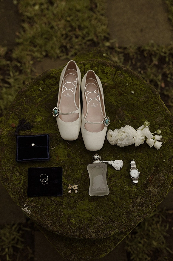 Wedding details flat lay with bridal shoes flat lay, rings in velvet boxes, perfume, watch and earrings on a mossy stone surface with greenery
