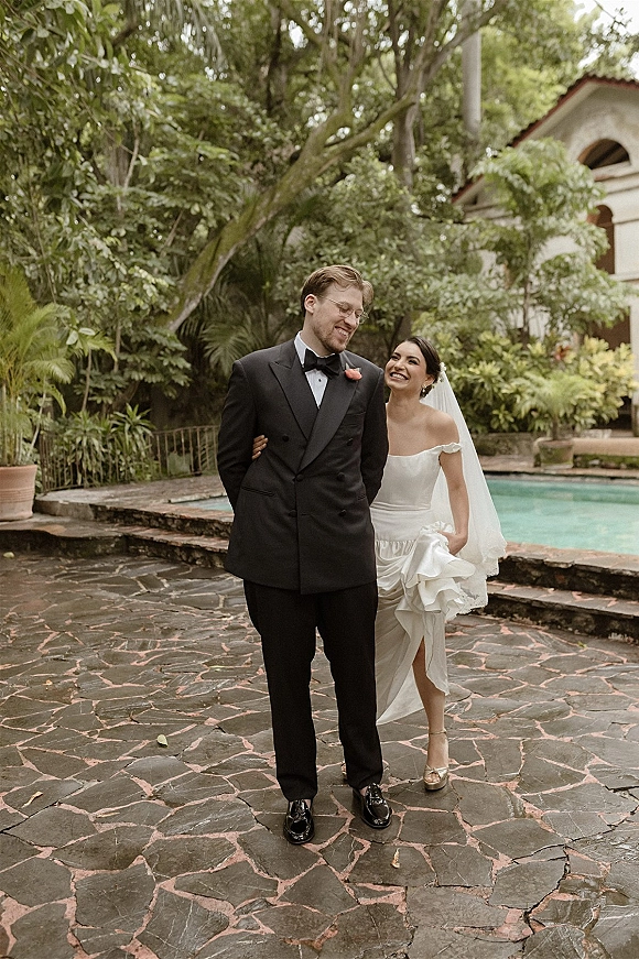 Couple portrait of bride and groom walking and laughing by a poolside stone patio, bride in veil and ruffle dress, groom in tuxedo