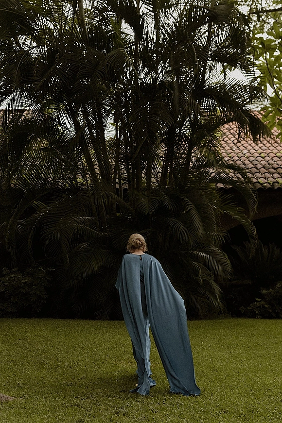 Wedding guest outfit in a blue cape dress with sleek low bun, seen from behind in a tropical courtyard with palm trees and terracotta roof
