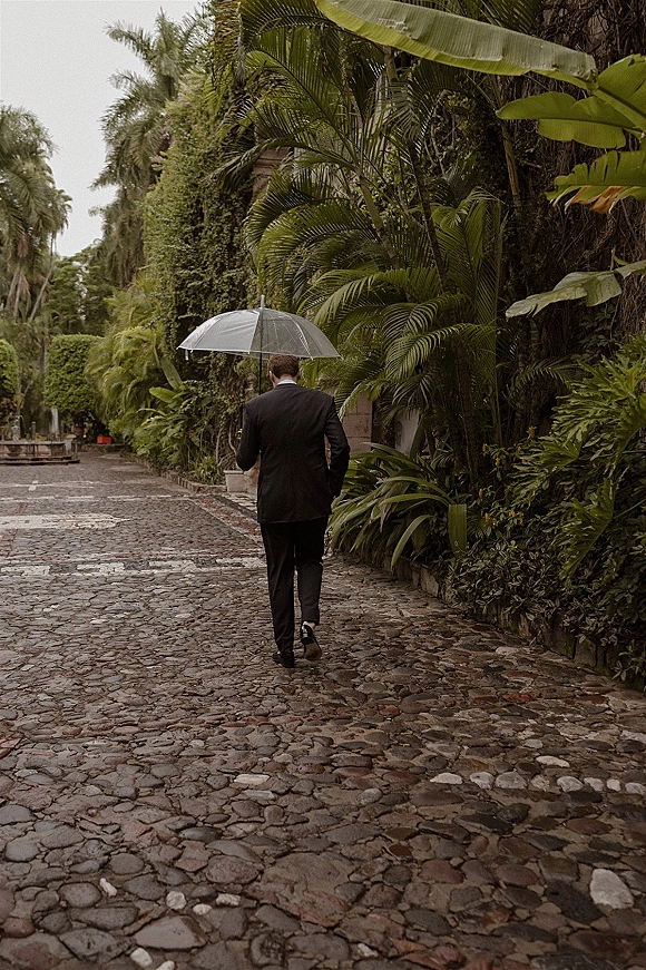Groom portrait of a man in a black suit walking away with an umbrella on a cobblestone garden walkway lined with palm trees