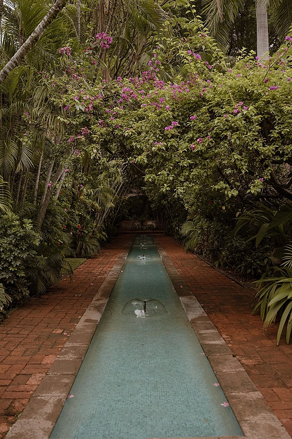 Garden walkway with tropical garden path lined by brick pavers and a narrow water channel, fountain jets, and bougainvillea amid palms