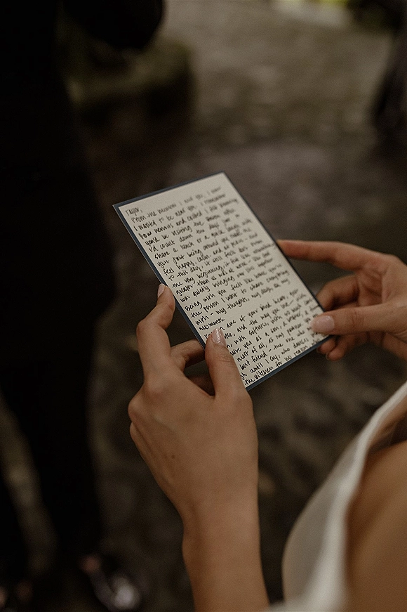 Wedding vows on a handwritten vow card held in the bride’s hands, dress strap visible, with blurred guests on a stone floor backdrop