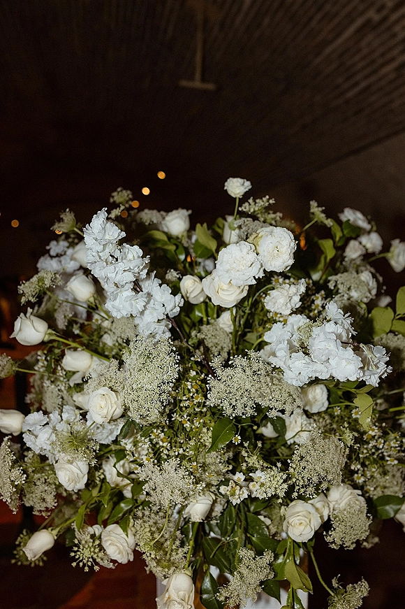 Wedding floral centerpiece with white rose centerpiece blooms, Queen Anne’s lace and greenery under string lights in a dark wood-ceiling venue