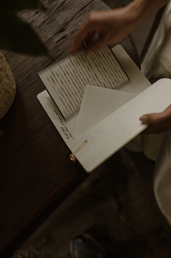 Wedding vows book open on a rustic wood table with handwritten wedding vows, a white envelope, note card, and paper clip held in hands