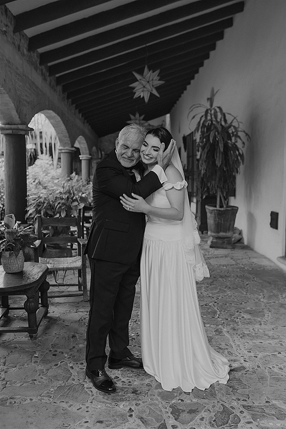 Father daughter wedding photo of the bride hugging her dad, veil trailing over an off the shoulder gown under star lanterns in an arched corridor